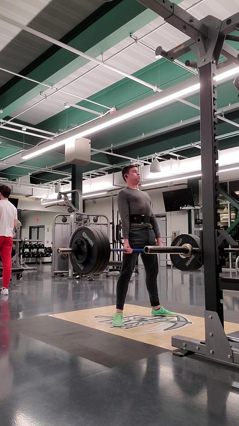 Duncan Bannon mid-lift with a loaded barbell in a university weight room.