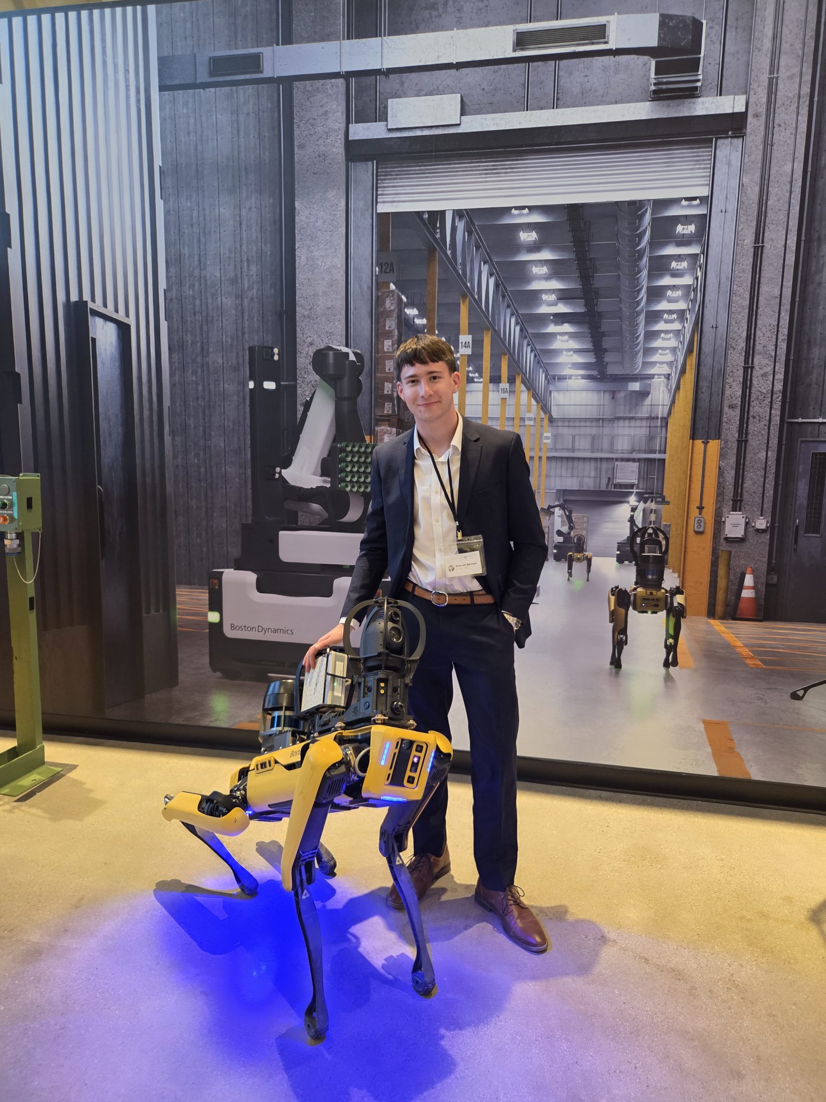 Duncan Bannon in a suit, standing beside a Boston Dynamics Spot robot on a facility floor.